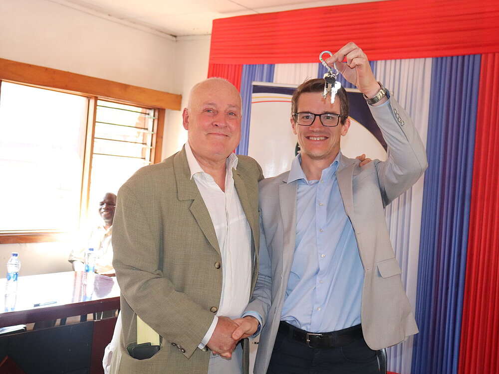 Two men smile at the camera and shake hands. One of them holds a key over his head.