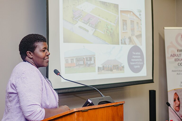 A woman stands at a wooden lectern and speaks into a microphone. She is wearing a light, lavender-coloured jacket. In the background, a presentation is projected onto a screen showing images of buildings and plans. On the right is a banner with a text and a photo. The atmosphere is professional and focussed.