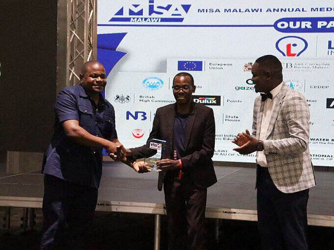 Three men on stage at the MISA Malawi Annual Media Awards, one hands over an award to another, the third applauds