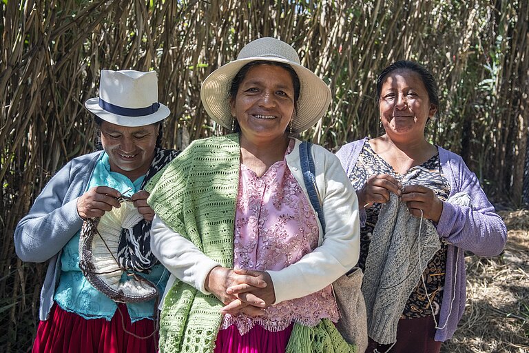 Drei Frauen stehen im Freien vor hohen, dichten Pflanzen. Die Frau in der Mitte lächelt freundlich in die Kamera. Die beiden Frauen an ihrer Seite konzentrieren sich auf ihr Handwerk – beide stricken etwas.