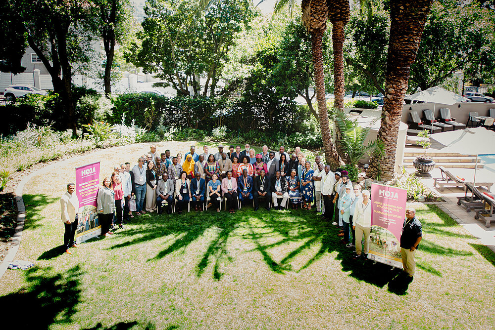 A group photo of people gathered on a lawn with palm trees. Two banners on the sides display the text 'MOJA: Adult Learning and Education for Sustainability and Resilience', and sunlight casts shadows across the grass.
