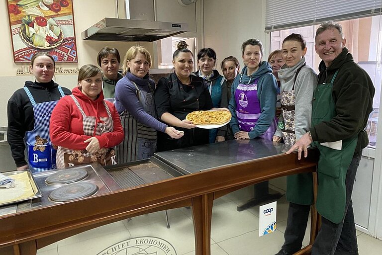 Zehn Frauen und ein Mann stehen in einer großen Küche, sie haben Kochschürzen um und präsentieren stolz eine fertige Pizza.