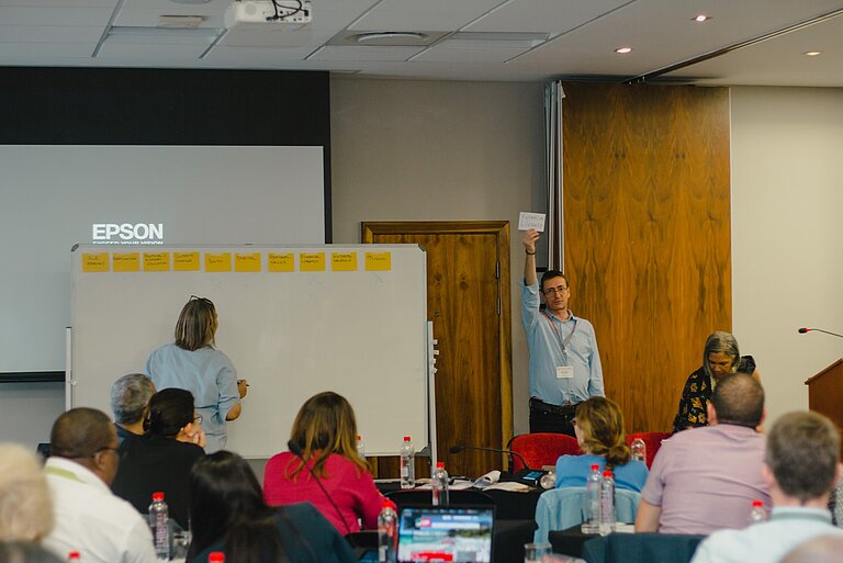 A man stands in front of a group of people who are sitting in front of him and listening to him. The man is holding a piece of paper in the air. Next to him, a woman stands facing a whiteboard on which similar notes are pinned. 