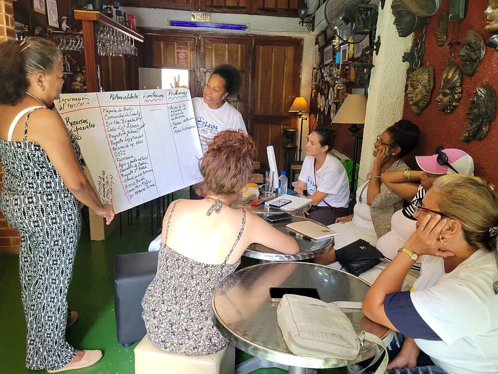 Two women present a self-labelled poster to a few other women. There is a bar in the background. The room is small, the women sit at silver round tables, close together and close to the presenters. Everyone is smiling and seems to be enjoying the exchange. 