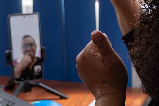 Im Vordergrund sind zwei erhoben Daumen zu sehen, die einer Person im Hintergrund gewidmet sind. Die Person ist via Videoanruf auf einem Smartphone zu sehen.