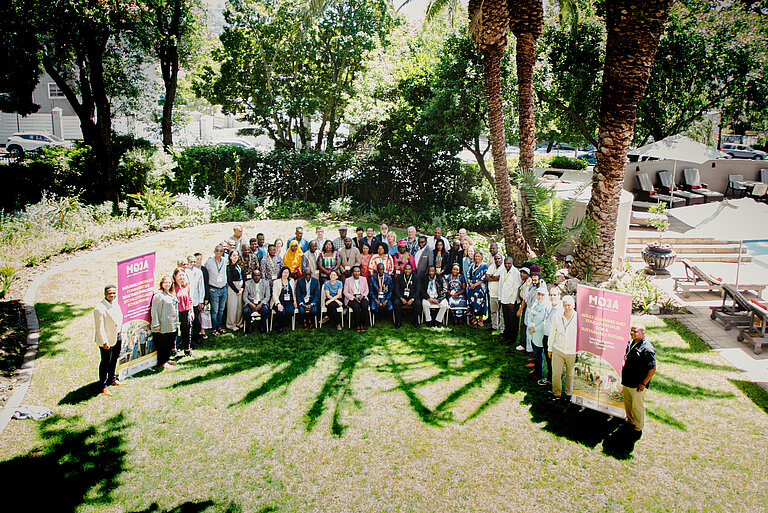 Ein Gruppenfoto von Menschen, die sich auf einem Rasen mit Palmen versammelt haben. Zwei Banner an den Seiten zeigen den Text „MOJA: Adult Learning and Education for Sustainability and Resilience“ (Erwachsenenbildung für Nachhaltigkeit und Resilienz), und das Sonnenlicht wirft Schatten auf das Gras.