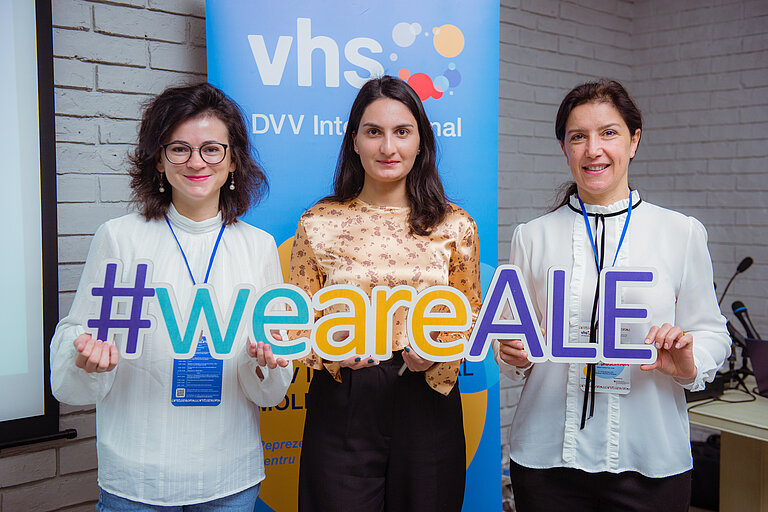 Drei Frauen stehen vor einem blauen Banner mit der Aufschrift "vhs DVV International" und halten gemeinsam ein großes Schild mit dem Hashtag "#weareALE". Sie lächeln und tragen Namensschilder an blauen Bändern. Die Umgebung hat eine moderne, helle Backsteinwand, und im Hintergrund befindet sich technisches Equipment. 
