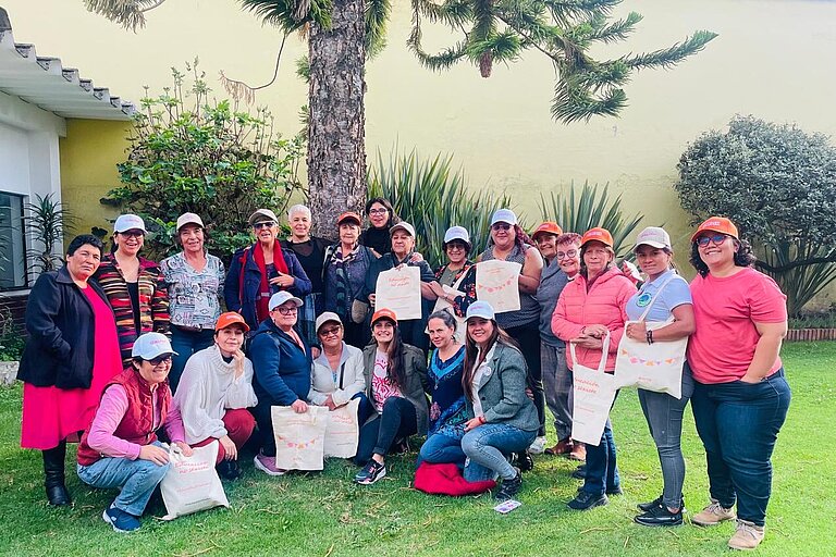  Eine Gruppe von Frauen unterschiedlichen Alters posiert gemeinsam im Freien auf einer Grünfläche vor einem Baum und einer gelben Hauswand. Viele von ihnen tragen Kappen in verschiedenen Farben und halten Stofftaschen mit Aufschriften. Sie lächeln , einige hocken oder knien vorne, während andere stehen. Die Umgebung wirkt freundlich und einladend mit Pflanzen und Sträuchern im Hintergrund.