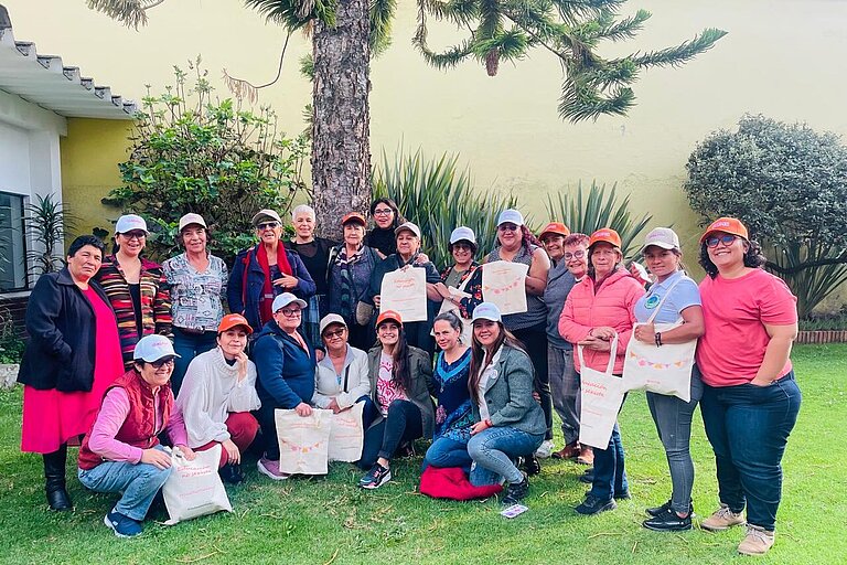  A group of women of different ages pose together outdoors on a green area in front of a tree and a yellow wall. Many of them are wearing caps in different colours and holding cloth bags with inscriptions. They are smiling, some are squatting or kneeling in front, while others are standing. The environment is friendly and inviting with plants and shrubs in the background.