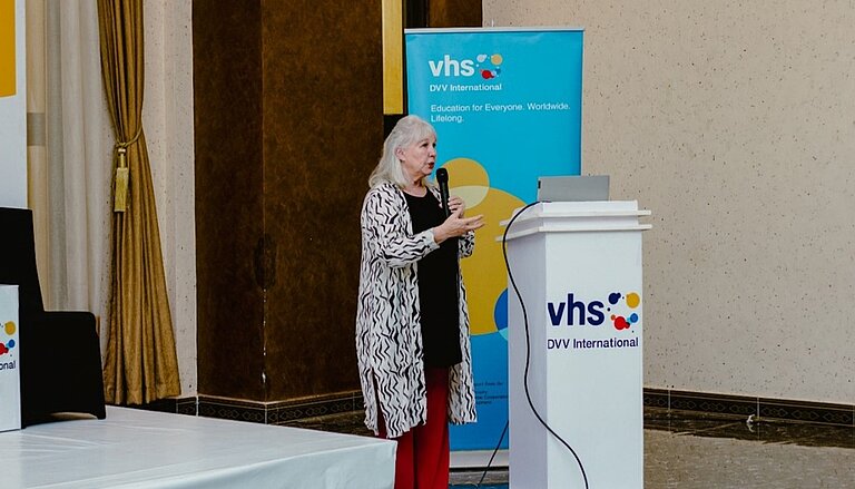 A woman stands at a lectern, speaking into a microphone while gesturing. She is in a formal event setting, with a banner displaying the DVV International logo positioned behind her.