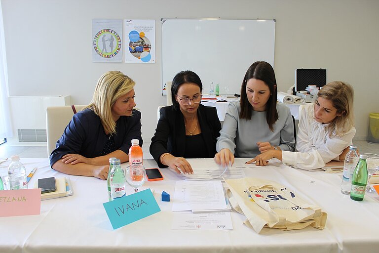 Vier Frauen sitzen an einem Tisch in einem Konferenzraum und arbeiten gemeinsam an den vor ihnen ausgebreiteten Dokumenten. Ihr Blick ist auf die Papiere gerichtet, und sie scheinen in eine Diskussion vertieft zu sein. 