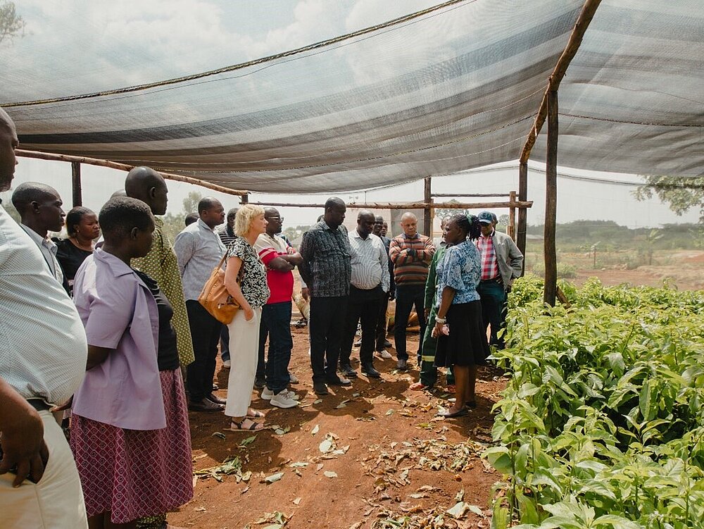  A group of people stand under a canopy in a field and listen to a woman. 