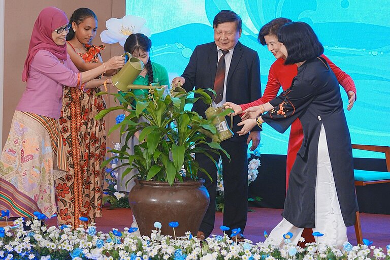 A group of six people, including women in traditional dress and a man in a suit, water a plant together in a large flower pot on a festively decorated stage. The background shows a blue digital representation of water waves. The stage is decorated with red fabrics, white and blue flowers and green plants.