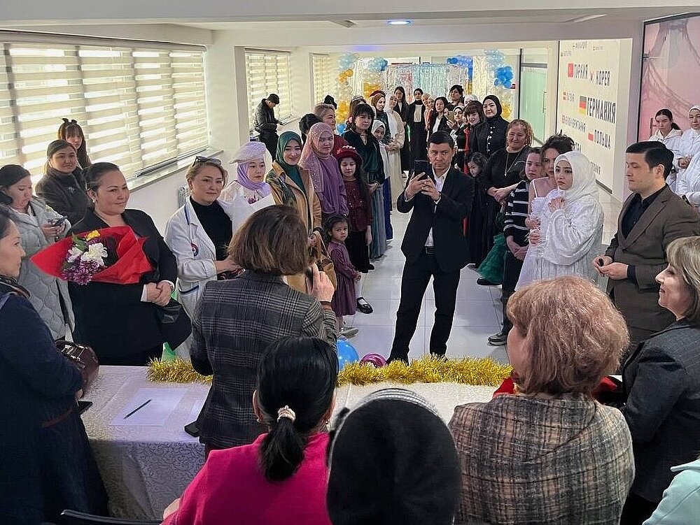 A group of people are standing in a brightly lit room. In the foreground, a woman is speaking to the others with her back to the camera. Next to her is a woman holding a large bouquet of flowers. Many of those present are wearing festive clothing, some are taking photos with their smartphones.