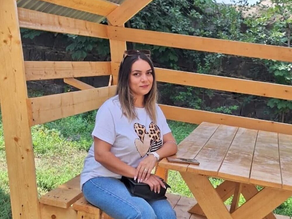 A woman is sitting outside on a bench at a table, holding her handbag on her lap.