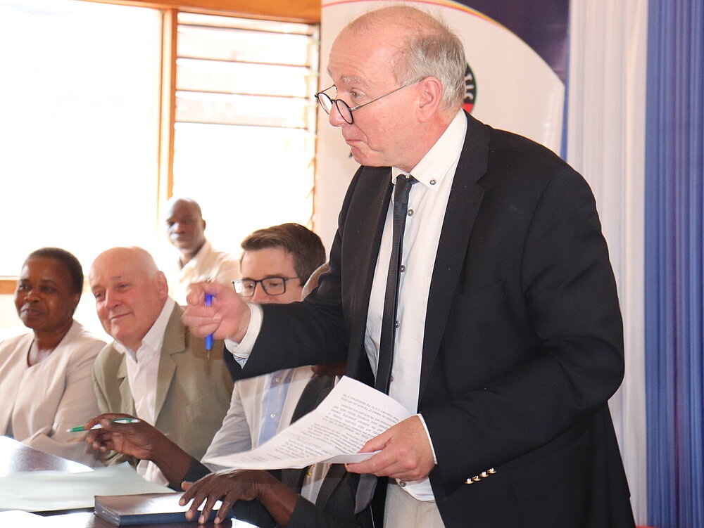 A man stands with a sheet of paper and a pen in his hands. Four other people can be seen sitting in the background.