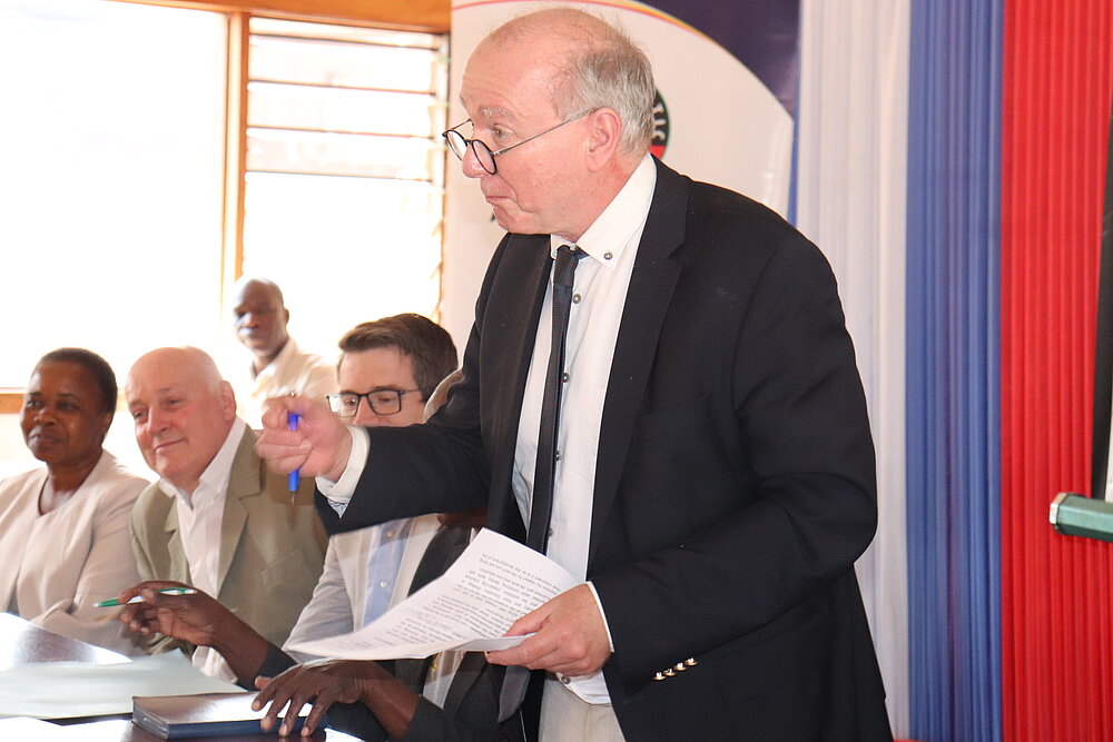 A man stands with a sheet of paper and a pen in his hands. Four other people can be seen sitting in the background.