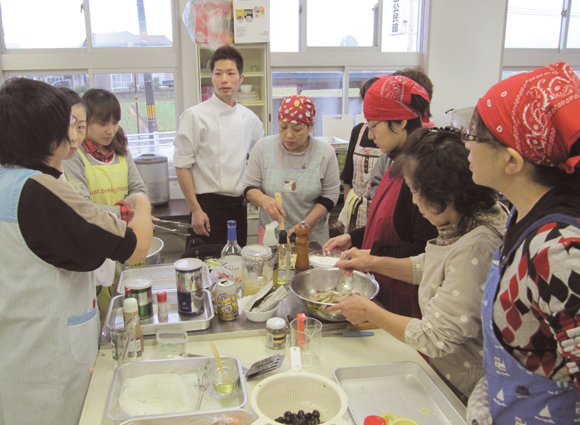 Course participants stand around a cooking island and handle various ingredients and utensils while the instructor stands by. 