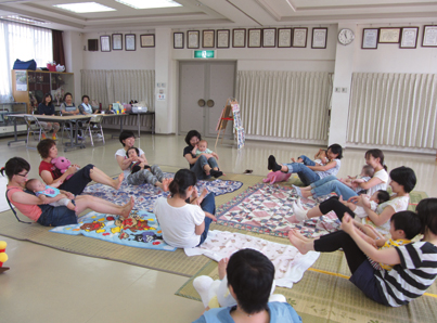 Mothers sit in a circle on mats on the floor with their babies on their laps and perform exercises. In the background, some women sit at a table and watch. 