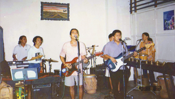 Musicians in a band play various instruments, such as electric guitar, bass and keyboard. Some also sing into a microphone. The band is surrounded by other instruments, such as a drum kit and other drums as well as music stands.