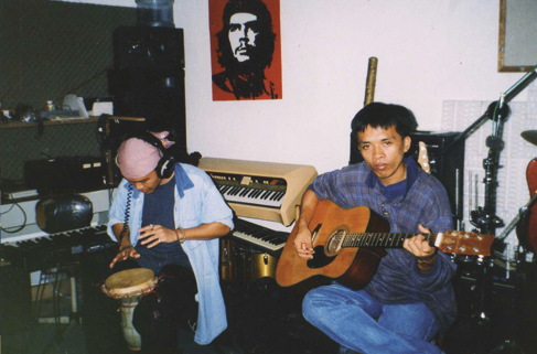 Two men are sitting in a recording room with various instruments. One of the men is playing the guitar and looking into the camera. The man next to him is drumming and has his headphones out.