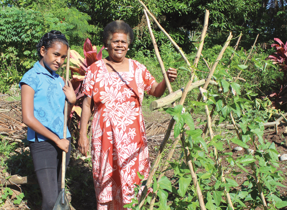 An older woman and a young girl are standing next to each other in a garden, next to a climbing plant. The girl has a rake in her hand, both are smiling into the camera. 