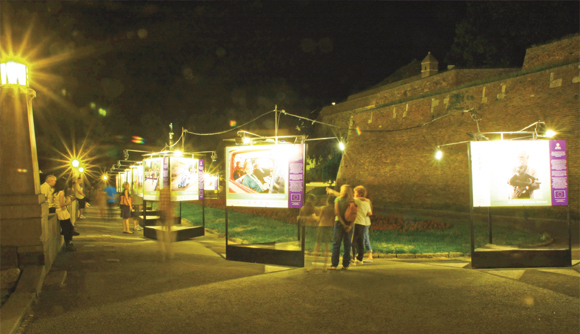 Une exposition photographique nocturne en plein air avec des panneaux lumineux installés le long d'un sentier pédestre. Les visiteurs regardent les photos tandis que des lanternes et des guirlandes lumineuses créent un éclairage d'ambiance. À l'arrière-plan, on aperçoit le mur d'une forteresse historique.