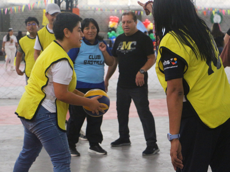 A group of people are taking part in a sporting activity in a hall. In the foreground, one person is holding a volleyball while another person gives instructions. There are other participants in the background.
