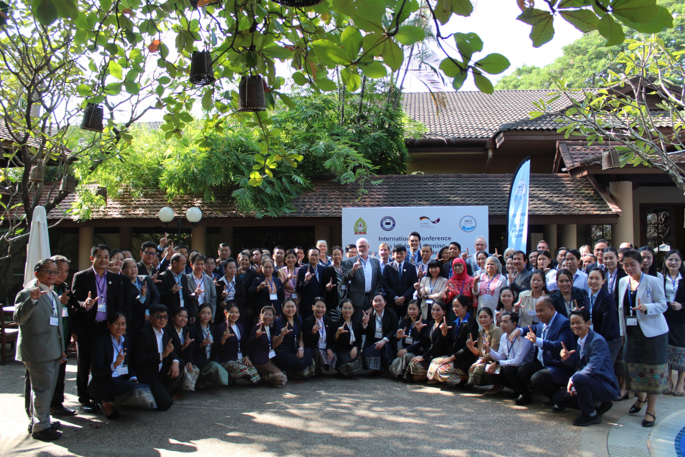 A large group of people pose together for an outdoor photo in front of a building with a traditional roof. In the background are green trees and plants. The group consists of men and women, mostly in formal dress, many wearing name tags. A banner in the background indicates an international conference.