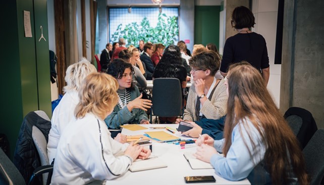 Several people are sitting at a table in a lively group discussion, they have notebooks, pens and documents in front of them. There are other groups in the background who are also taking part in discussions.