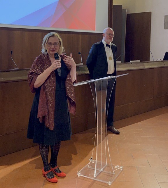 A woman stands at a lectern and speaks into a microphone. She is wearing a black dress and a reddish-brown stole. In the background, a man in a suit with a gold medal around his neck stands in front of wooden panelling. At the top of the picture is a screen with a graphic image.